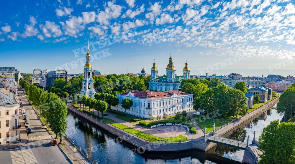 На этой фотографии представлен панорамный вид на Никольский морской собор в Санкт-Петербурге. Видны его яркие золотые купола и звонница, отражающиеся в спокойной воде канала. Эта красочная сцена, залитая солнечным светом и украшенная ясным небом с пушистыми облаками, может быть использована в туристических брошюрах, образовательных материалах о русской архитектуре или как элемент декора.